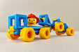A vintage toy construction vehicle set made of bright blue and yellow plastic pieces, featuring a smiling yellow face driver wearing a red hard hat, photographed on a neutral light beige background.