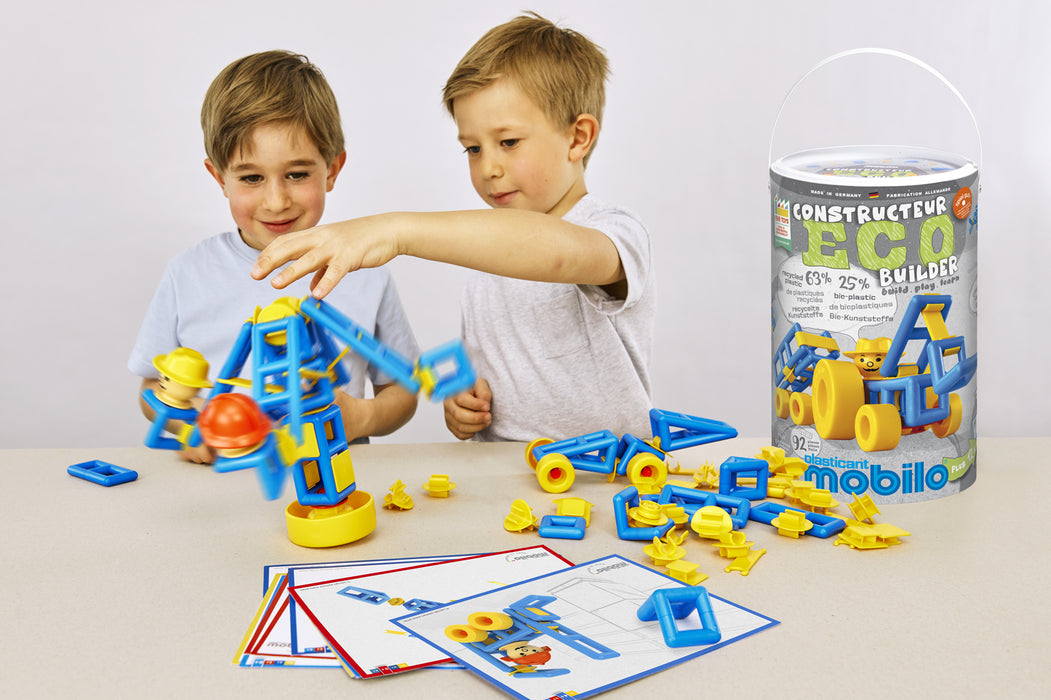 Two young boys play with a colorful mobile construction toy set on a light table, with loose blue and yellow building blocks scattered around and the product canister standing on the right.
