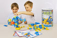 Two young boys play with a colorful mobile construction toy set on a light table, with loose blue and yellow building blocks scattered around and the product canister standing on the right.