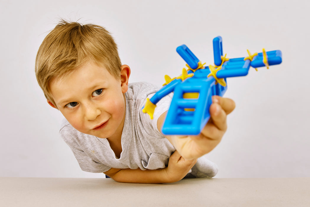 A young boy with light brown hair lies on his stomach, holding up a blue plastic toy with yellow elastic bands against a plain light gray background.